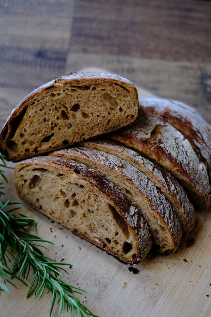 Close-up of a sliced, crusty sourdough loaf with fresh rosemary on a wooden surface.